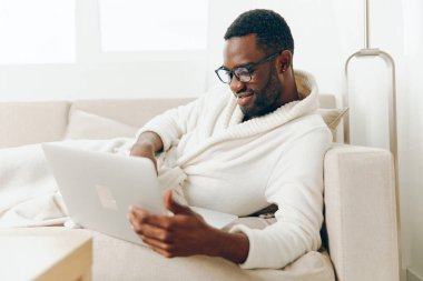 Modern bir ev ofisinde dizüstü bilgisayarıyla çalışan Afrikalı Amerikalı serbest çalışan, yüzünde bir gülümsemeyle, rahat bir şekilde kanepede oturan, kendini suya atarken huzurlu bir sabah.