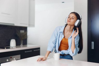 Kadın kulaklıkla müzik dinlerken modern mutfakta tezgahın üzerinde duruyor, evde dinlenme ve rahatlama kavramı
