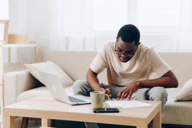 Genç Afrikalı Amerikalı serbest çalışan, rahat oturma odasındaki modern bir kanepede otururken yüzünde bir gülümsemeyle online eğitimine odaklanmış,