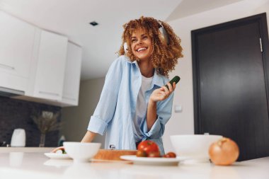 Kıvırcık saçlı, mutlu bir kadın elinde bir tabak yemekle mutfakta duruyor, gülümsüyor ve yemeğin tadını çıkarıyor.