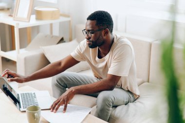 Modern İçişleri Bakanlığı 'nda dizüstü bilgisayarda çalışan gülümseyen Afrikalı Amerikalı Adam gündelik giysiler içinde oturma odasındaki rahat siyah bir kanepede oturuyor.