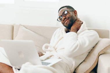 Gülümseyen Afrikalı Amerikalı Freelancer, Bornoz 'da dizüstü bilgisayarda çalışıyor, Modern İçişleri Bakanlığı' nda koltukta oturuyor genç adam bilgisayarında daktilo yazıyor, verimli bir sabahın keyfini çıkarıyor.