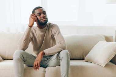Başı ağrıyan yorgun Afrikalı Amerikalı bir adam, evdeki kanepede tek başına oturuyor. Stresli ve hayal kırıklığına uğramış görünüyor. Yüzünde acı ve rahatsızlık belirtileri var.