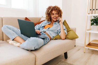 Gülümseyen kadın rahat oturma odasında dizüstü bilgisayarda çalışıyor, teknolojiyle bağlantı kuruyor.