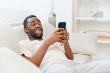 Modern dairesinde siyah bir koltukta oturan mutlu Afrikalı Amerikalı adam, cep telefonunu sohbet etmek ve mesajlar yazmak için kullanıyor. Kablosuz iletişimin keyfini çıkarırken kendine güven ve rahatlama yayıyor.