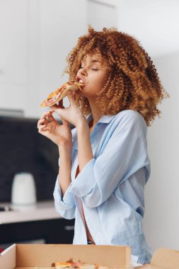 Kıvırcık saçlı genç bir kadın evde açık pizza kutusunun önünde pizza yiyor.