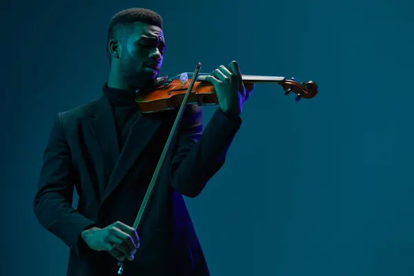 スタジオの設定で活気に満ちたブルーの背景に対してバイオリンを実行する黒いスーツのエレガントな男 ロイヤリティフリーのストック写真