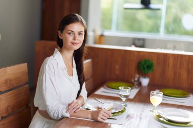 Romantik Akşam Yemeği Randevusu. Esmer saçlı, sıradan giyinmiş güzel bir kadın, rahat bir yemek odasında şık bir yemek masasında oturur. Masa zarif tabaklar ve güzel bir tabakla hazırdır.