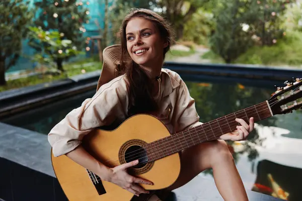 A woman is sitting on a stone wall with an acoustic guitar in front of a pond