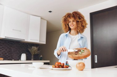 Kıvırcık saçlı bir kadın mutfaktaki tahta kesme tahtasının üzerinde taze sebzeler hazırlıyor.