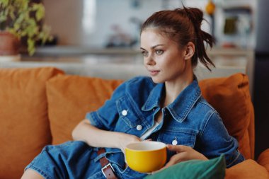 Rahat bir kadın, elinde kahve fincanıyla kanepede oturmuş, sıcak ev ortamında kameraya bakıyor.