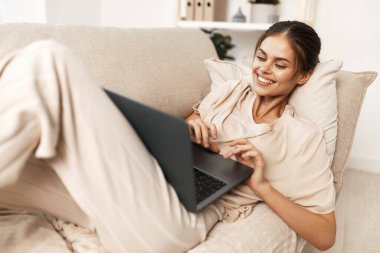 Evde dizüstü bilgisayar kullanan, modern oturma odasındaki koltukta oturan gülümseyen kadın..