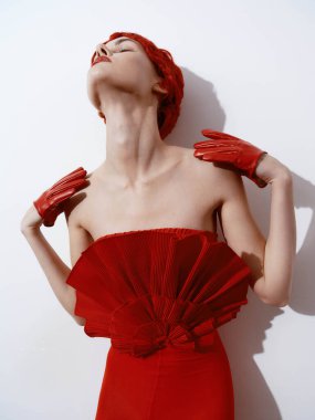 Kırmızı elbiseli ve eldivenli zarif bir kadın ellerini beyaz duvara dayayıp poz veriyor.