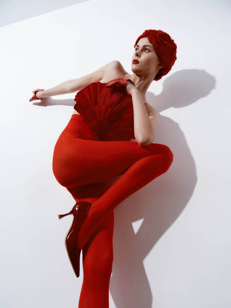 Stylish woman in a red dress and high heels posing against a white wall with a shadow