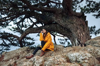 Doğada sakin bir an. Sarı paltolu bir kadın. Büyük bir kayanın üzerinde bir ağacın yanında oturuyor.