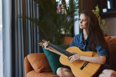 Pencerenin önündeki koltukta akustik gitar çalan kadın.