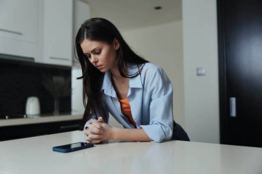 Mutfak tezgahında oturan mavi gömlekli bir kadın telefona bakıyor, günlük yaşam tarzı konsepti.