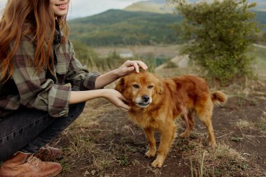 Huzurlu bir kadın, sakin bir tepede, arkası görkemli dağlarla kaplı yeşil bir tarlaya bakan dost canlısı bir evcil köpekle eğleniyor.