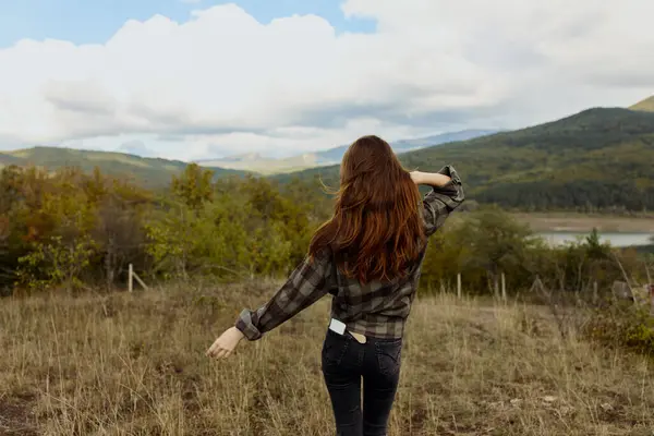 stock image A woman enjoying the serene beauty of nature in a plaid shirt standing in a picturesque field with majestic mountains in the background