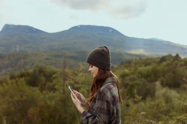 Şık şapkalı ve ekose gömlekli şık bir kadın akıllı telefonunu kullanarak doğanın huzurlu güzelliklerini keşfediyor.