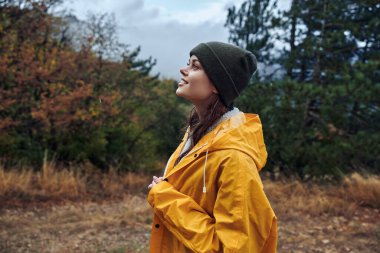 Sarı yağmurluklu genç bir kadın sakin bir alanda duruyor doğa güzelliği ve seyahat konseptiyle çevrili.