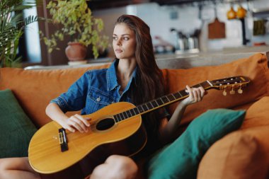 Oturma odasındaki bir bitkinin önünde akustik gitarla kanepede oturan genç bir kadın.