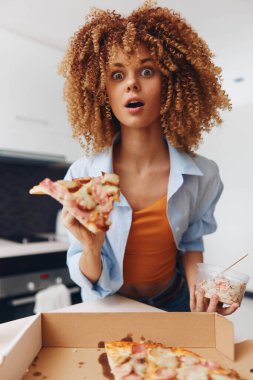 Kıvırcık saçlı genç bir kadın karton bir kutunun önünde pizza yiyor.