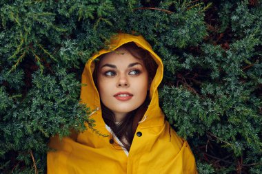 Sarı yağmurluklu genç bir kadın yemyeşil ağaçlar ve çalılarla çevrili doğanın tadını çıkarıyor.