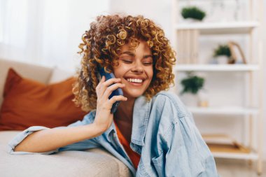 Evdeki neşeli kadın, rahat koltukta oturuyor elinde bir cep telefonu, gülümsüyor ve online oyunlardan zevk alıyor.