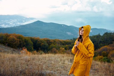 Sarı yağmurluklu kadın huzur dolu bir yalnızlık içinde tepede dururken görkemli dağlara hayran.