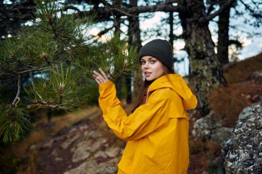 Bir doğa gezisi sırasında ormanda çam ağaçlarının arasında duran sarı yağmurluklu genç bir kadın.