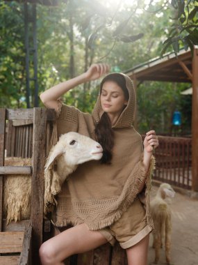 Tahta bir bankta oturan bir kadın önündeki koyunla kahverengi bir panço giyiyor.