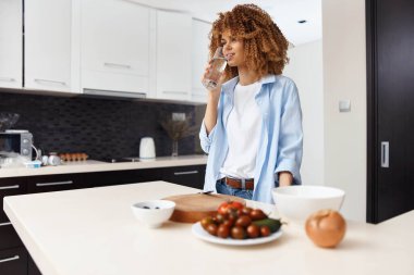 Çekici Afrikalı Amerikalı kadın evdeki modern mutfağında ferahlatıcı bir bardak su içiyor.