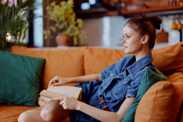 Woman relaxing on couch at home, reading book in denim shirt