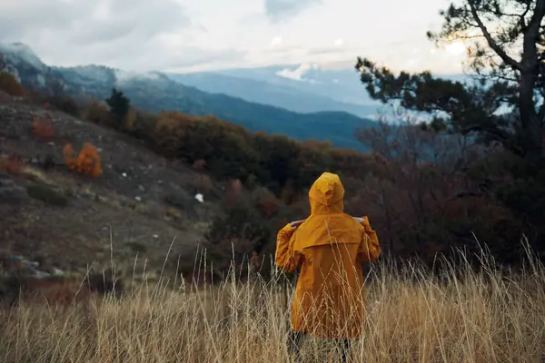 stock image A person in a yellow raincoat standing in a picturesque field with majestic mountains in the background during a travel adventure