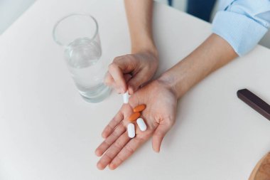 Arka planda bıçak ve su bardağı olan beyaz masanın üzerinde ilaç ve bardak tutan eller.