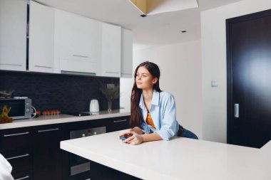 Kadın mutfak tezgahında elinde uzaktan kumandayla oturmuş televizyon izliyor ve dinleniyor.