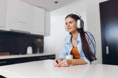 Mutfak masasında kulaklıkla müzik dinlerken bir fincan kahvenin tadını çıkaran genç bir kadın.