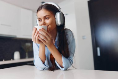 Genç bir kadın, modern bir mutfak ortamında kulaklıklar ve kahveyle sessiz bir sabahın tadını çıkarıyor.