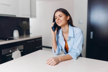 Rahat bir ev ortamında doğal ışıklandırmayla mutfak masasında akıllı bir telefonla sohbet eden bir kadın.