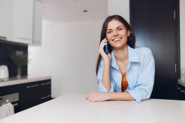 Mutfak masasındaki mutlu kadın cep telefonuyla konuşuyor, kameraya gülümsüyor, ev ortamında.