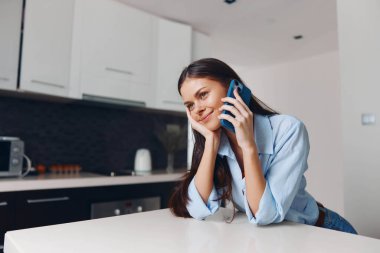 Kadın mutfak tezgahında oturmuş cep telefonuyla konuşuyor ve günlük ev ortamında kameraya bakıyor.