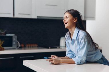 Mutfak tezgahında oturan mutlu kadın cep telefonu elinde, gülümsüyor.