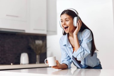 Genç bir kadın, mutfaktaki rahat atmosferde kulaklık ve kahveyle müzik dinlemekten hoşlanıyor.