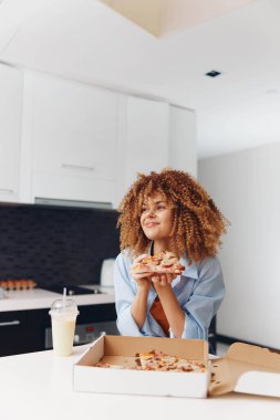 Kıvırcık saçlı bir kadın masada iki dilim pizzanın tadını çıkarıyor.