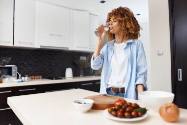 Genç Afrikalı Amerikalı kadın evdeki modern mutfağında susuz kalıp ferahlatıcı bir bardak suyun tadını çıkarıyor.