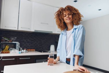 Afrika kökenli Amerikalı bir kadın elinde bir bardak suyla modern mutfakta duruyor. Evinde güzel ve sağlıklı görünüyor.