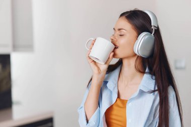 Mutfakta kulaklıklı bir kadın kahve içiyor ve telefonda müzik dinliyor.