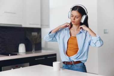 Mutfakta kulaklıkla müzik dinlerken sabah kahve molası veren genç bir kadın.