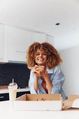 Mutlu Afrikalı Amerikalı kadın sıcak mutfakta lezzetli pizza ve sütün tadını çıkarıyor.